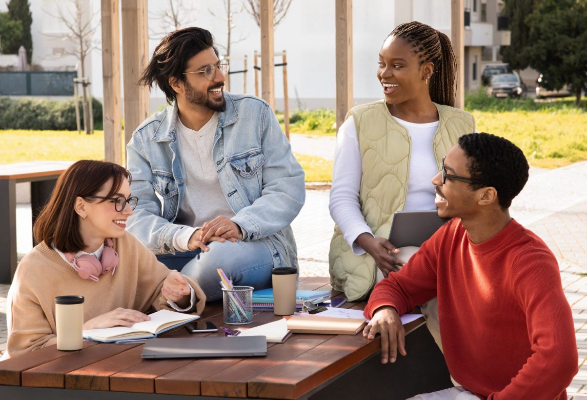 Cheerful international millennial students study and prepare project together at table with gadgets in campus outdoor. Coffee break and education, knowledge and teamwork, rest and communication